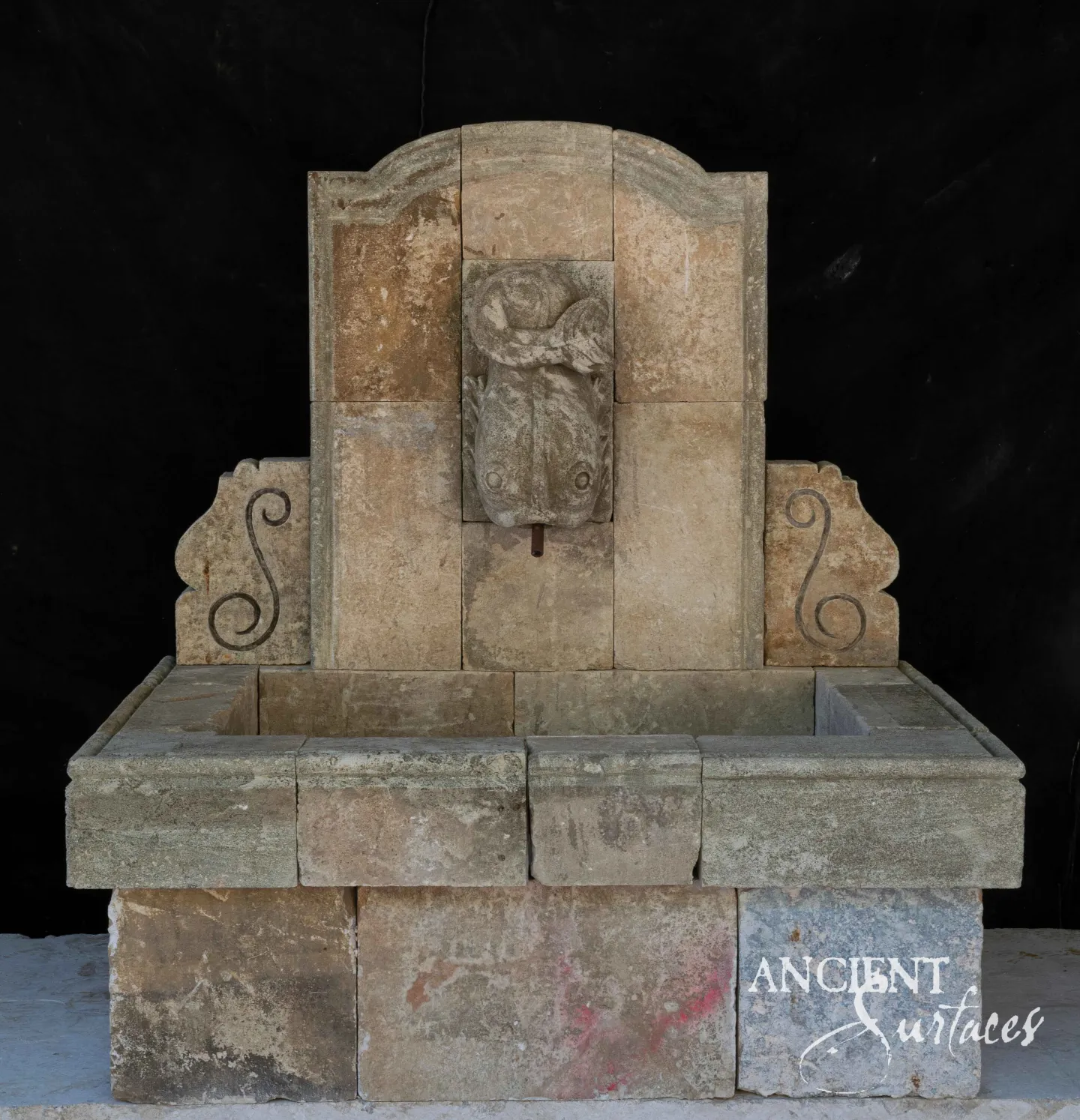 Reclaimed limestone wall fountain with a carved dolphin-mask spout and iron scroll brackets over a stone trough