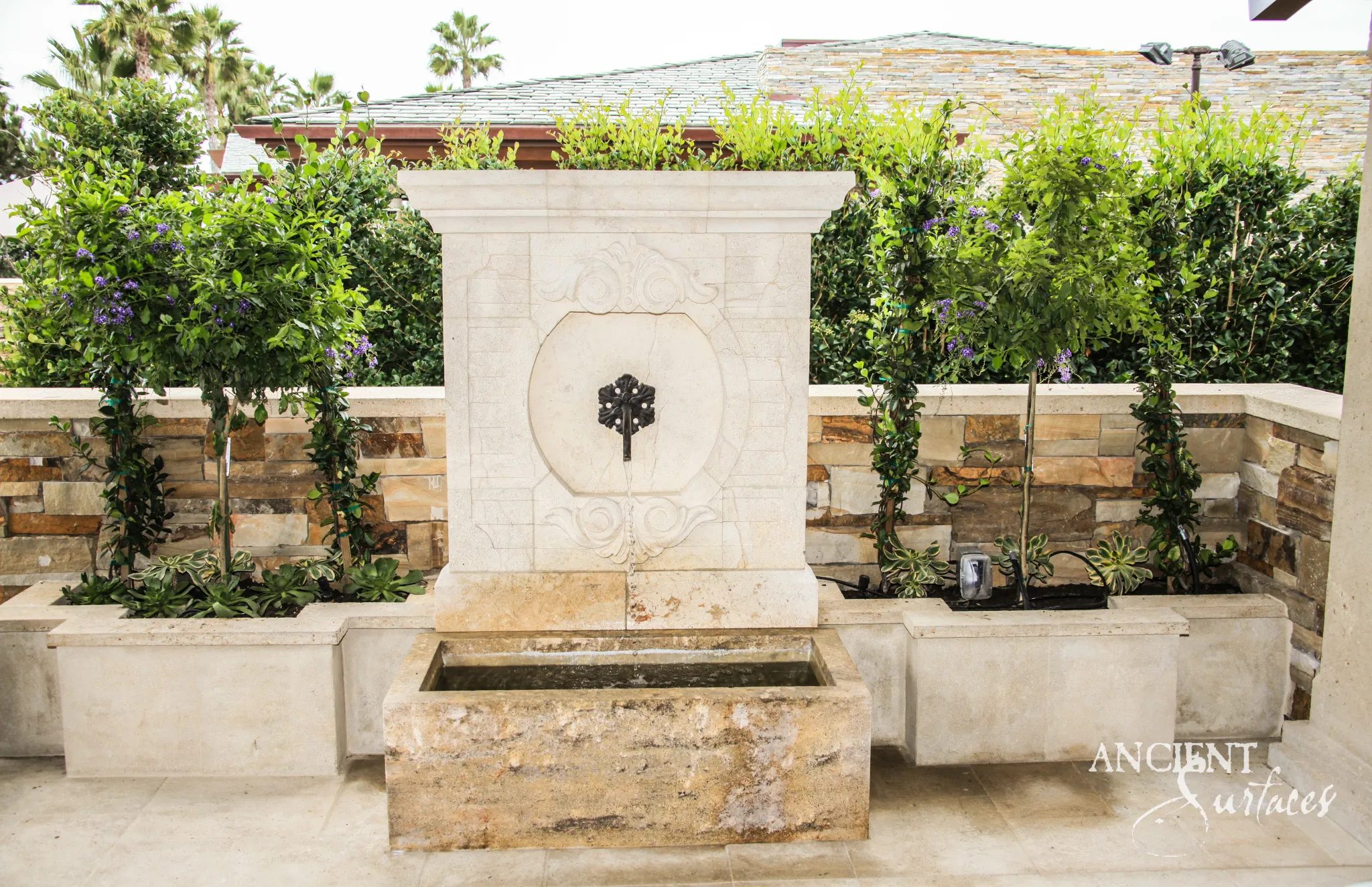 Reclaimed limestone wall fountain with a carved panel and iron rosette spout installed on a formal planted terrace